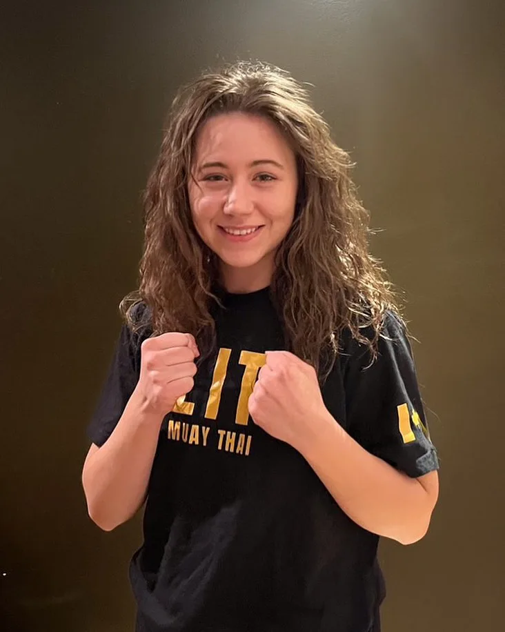 A smiling individual stands confidently with fists raised, wearing a black Muay Thai shirt
