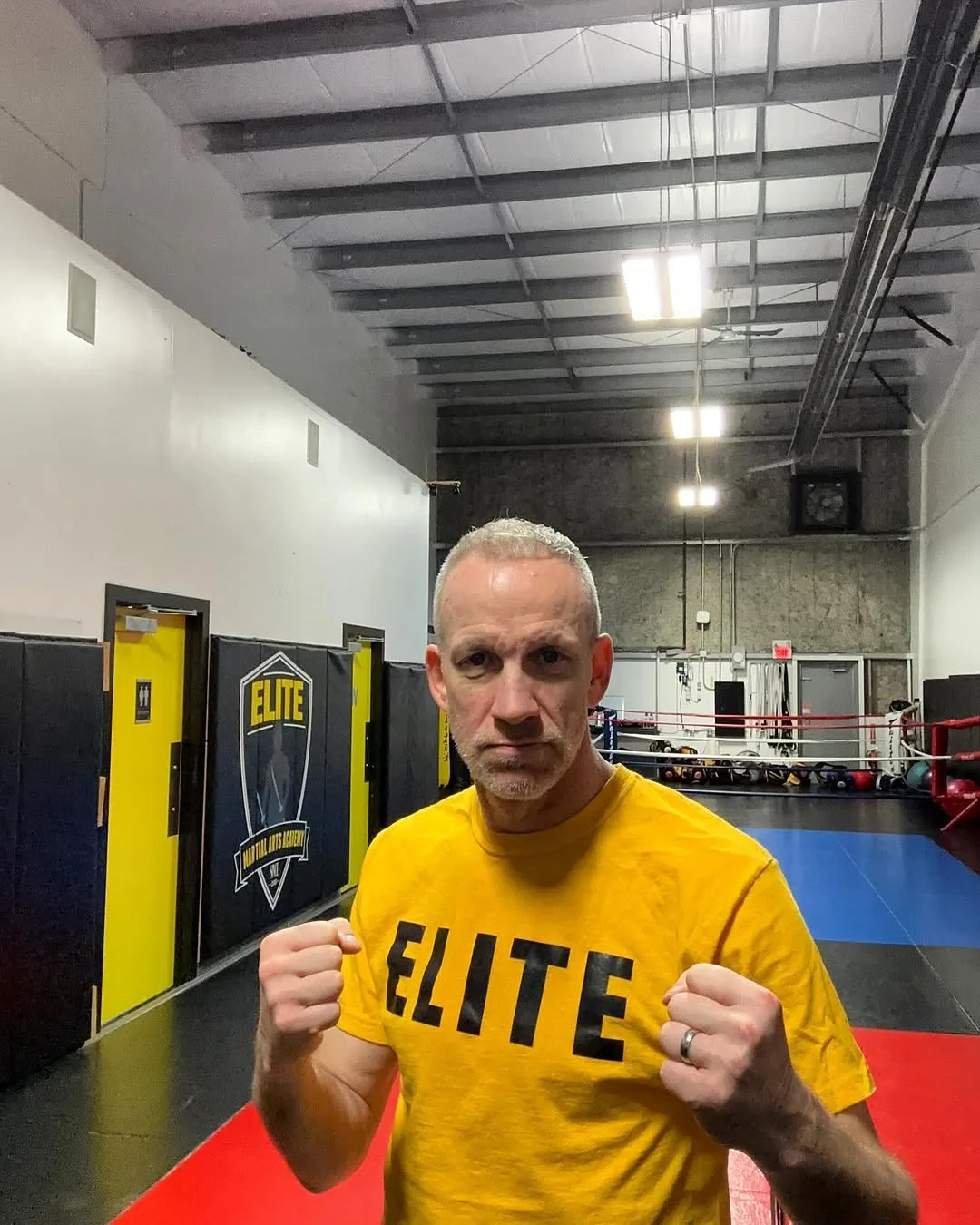 A man in a yellow shirt poses with fists raised in a boxing gym