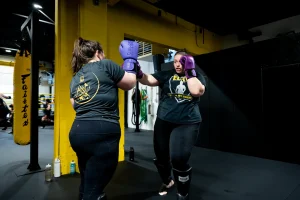 Two individuals spar in a gym, wearing protective gear and focused expressions